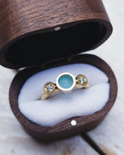Load image into Gallery viewer, Side view of an ocean-inspired CAVERN engagement ring showing the trilogy-style bezel setting that has luxurious blue sea glass and diamonds from the sea set within it.