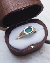 Load image into Gallery viewer, Close-up of a bespoke signet engagement ring showing handcarved oval teal sea glass in the center with twelve grain set ocean diamonds that taper down the freeform gold band. 

