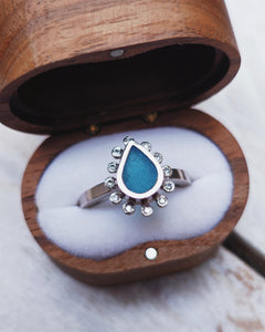 Side view of a Cornish handcrafted HALO engagement ring showcasing the bezel setting that features waterdrop-shaped blue sea glass encircled by a halo of twelve ocean diamonds.